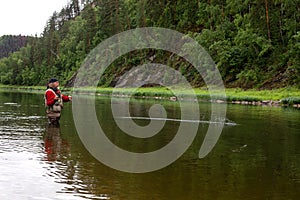 Side view portrait of brutal aged fisherman standing hip-deep in narrow forest river at the moment of catching fish by fishing