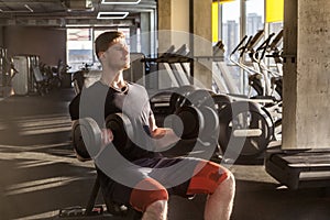 Side view portrait of concentration young adult man handsome athlete working out in gym, sitting on a bench and holding two