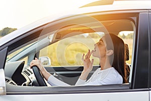 Side view portrait of dark haired beautiful woman driving a car and looking at mirror touching up her makeup, attractive female