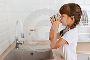 Side view portrait of dark haired little girl with pigtails wearing white t shirt holding cup and standing near the kitchen faucet