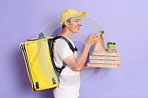 Side view portrait of delivery man in yellow cap and T-shirt with thermo backpack holding pizza in cardboard flatbox and takeaway