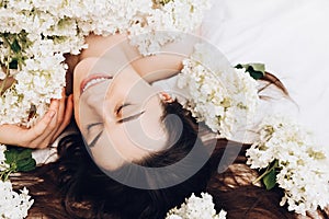 Side view portrait of dreamful beautiful young woman with closed eyes and sincere smile lying in cozy white bed, summer flowers