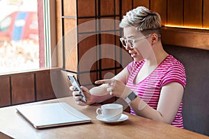 Side view portrait of excited young blogger with short hair in pink t-shirt is sitting in cafe, holding phone and making video