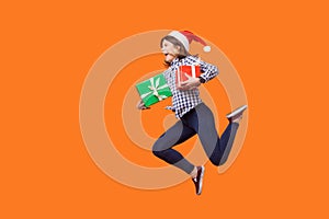 Side view portrait of extremely joyous brunette woman in santa hat jumping or running in air with wrapped xmas gift boxes,