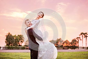 Side view portrait of happy bridegroom carrying bride on field during sunset