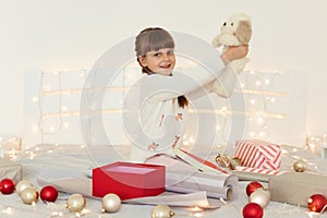 Side view portrait of little girl wearing white sweater holding soft toy dog, sitting on bed with Christmas decoration and garland