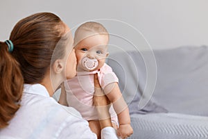 Side view portrait of lovely woman with dark hair woman playing with her infant daughter at home, mother kissing her baby,