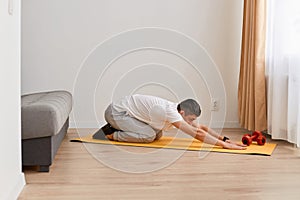 Side view portrait of man wearing sportswear doing sport exercises at home practicing yoga, doing seated forward bend Balasana