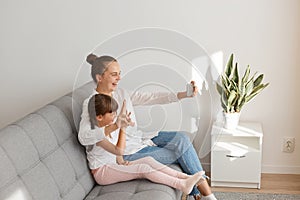 Side view portrait of mother and daughter taking selfie at home in the living room while sitting on sofa, broadcasting livestream