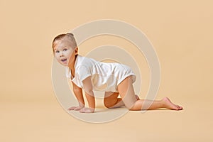 Side view portrait of pretty little baby, girl crawling across floor in cute white romper against pastel color