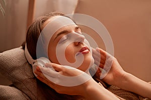 Side view portrait of relaxed young adult woman listening music while resting on couch at home, keeps eyes closed, touching her