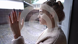 Side view portrait of sad brokenhearted teenage girl looking out the window on rainy day touching glass. Depressed