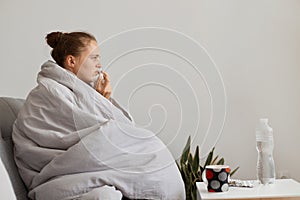 Side view portrait of sad sick woman with hair bun sitting on sofa wrapped in blanket, suffering runny nose and sneezing, using