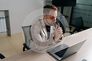 Side view portrait of serious pensive businessman in suit holding smartphone sitting at office desk with laptop computer