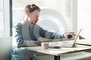 Side view portrait of serious young businessman in blue shirt are sitting and pointing at target on laptop dislpay with attention