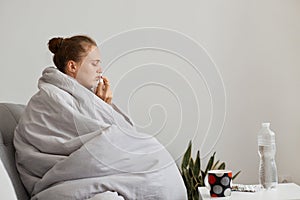 Side view portrait of sick upset woman with hair bun sitting on sofa wrapped in blanket using nasal spray, suffering runny nose