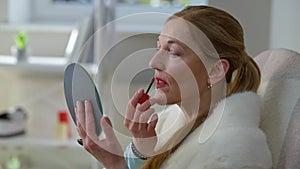 Side view portrait of slim middle aged Caucasian woman applying lipstick looking at hand mirror. Elegant wealthy rich