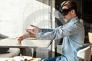 Side view portrait of suprised bearded young adult man in blue denim shirt sitting in cafe, wearing vr and watching video on