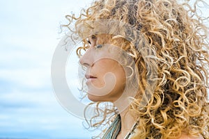 Side view portrait of woman with beautiful long blonde curly hair and eyes closed and thoughts expression. Blue sky background.