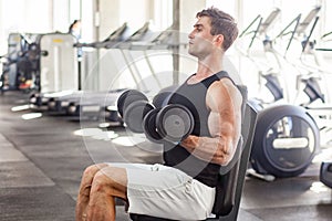 Side view portrait of young adult man muscular built handsome athlete working out in a gym, sitting on a bench and holding two