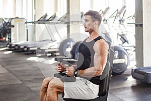 Side view portrait of young adult man muscular built handsome athlete working out in a gym, sitting on bench and holding two