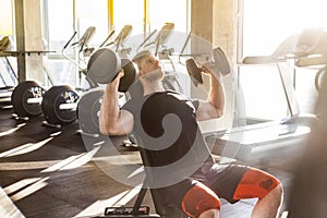 Side view portrait of young adult sport man training at gym alone. Athlete workout in gym, sitting and holding two dumbbell with