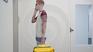 Side view portrait of young Caucasian man talking on phone waiting for lift with yellow food delivery bag. Courier