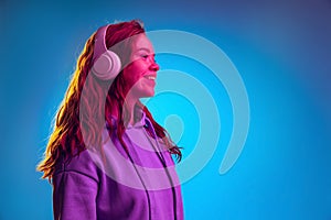 Side view portrait of young girl in white headphones posing isolated over blue studio background in pink neon light