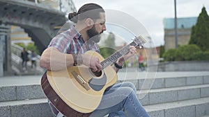 Side view portrait of young man playing guitar sitting on city stairs. Concentrated Caucasian guitarist performing alone