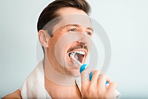 Side view portrait of young man with towel on shoulders brushing white teeth with modern electric toothbrush, close-up.
