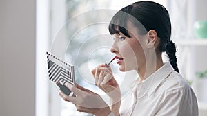 Side view portrait of young slim confident businesswoman applying lipstick in office. Beautiful Caucasian brunette woman