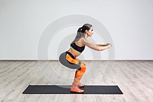 Side view portrait of young sporty healthy beautiful woman in black top and orange leggings doing squatting with elastic