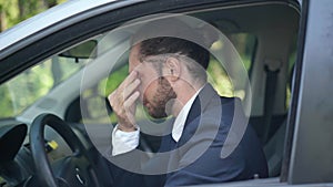 Side view portrait of young stressed Caucasian man in suit sitting on driver's seat rubbing temples sighing