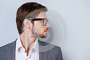 Side-view portrait of young successful serious man with bristle in glasses and wearing suit standing on gray background