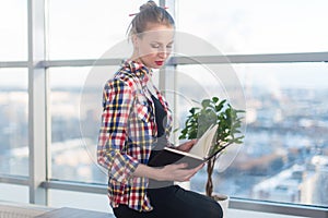 Side view portrait of young woman sitting, looking down, reading the book, learning at light room in morning with her