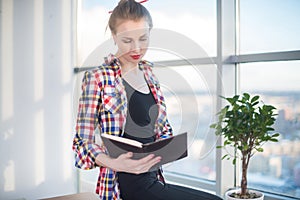 Side view portrait of young woman sitting, looking down, reading the book, learning at light room in morning with her