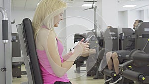 Side view of positive blond woman typing on smartphone with men training at the background. Portrait of young beautiful