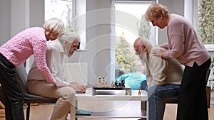 Side view positive senior wives supporting husbands playing chess indoors. Happy Caucasian women endorsing men enjoying