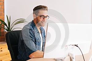 Side view of positive young designer male in stylish glasses working on desktop computer sitting at desk at home office