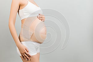 Side view of pregnant woman in underwear wearing elastic maternity band at gray background with copy space. Close up of orthopedic