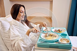 Side view of pregnant woman wearing bathrobe cutting meat during having lunch