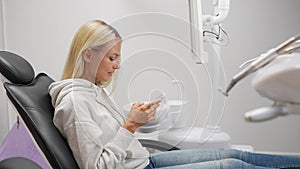 Side view of pretty blonde young woman sitting in dental chair using mobile phone while waiting for dentist in modern