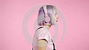 Side view. Pretty girl with dyed violet hair listening to music, smiling, dancing in headphones in studio against pink