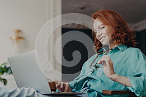 Side view of pretty redhead woman concentrated on remote job, prepares publication for web page, looks gladfully at laptop