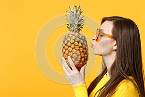 Side view of pretty young woman in heart glasses holding in hands kissing fresh ripe pineapple fruit isolated on yellow