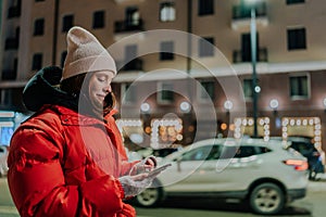 Side view of pretty young woman wearing hat and winter jacket calling taxi using smartphone app. Frozen smiling female