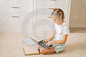 Side view of primary little child girl doing homework using laptop computer for distance education online sitting on