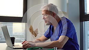 Side view of professional mature male doctor wearing blue uniform using laptop computer in office.