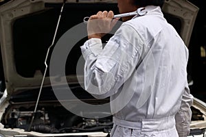 Side view of professional young mechanic man in uniform holding wrench against car in open hood at the repair garage.