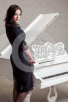 Side view profile of sensual brunette woman in lace black classic dress standing near white piano and looking at camera